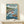 May include: Framed Studio Ghibli-style anime poster showing red-haired Ponyo emerging from ocean wave next to boy in wooden boat, with coastal village, lighthouse, and mountains in background. Japanese and English text at top and bottom. Displayed on light wood shelf with small white gift box, against cream wall.