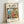 May include: Framed Studio Ghibli poster print featuring Ponyo character in red dress standing in cozy kitchen with ocean cliff view through window. Artwork shows detailed anime-style illustration with warm colors, wooden kitchen elements, and Japanese text above English title. Light wooden frame leans against white wall with soft natural lighting.