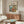 May include: Framed Studio Ghibli Ponyo poster mounted above wooden mid-century modern credenza in minimalist living room. Poster shows red-haired child character cooking in cozy kitchen overlooking ocean cliffs. Room features cream upholstered chair, glass-top oval coffee table, white table lamp, and warm natural lighting creating shadows on light walls.
