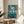 May include: Framed Ponyo movie poster displayed on round wooden side table next to beige upholstered chair. Poster features red-haired character emerging from swirling ocean water with coastal buildings and lighthouse in background. Underwater scene shows colorful sea life including fish and jellyfish. Japanese text at top, English title at bottom. Natural lighting creates soft shadows on white wall behind.