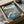 May include: Framed Studio Ghibli-style poster showing red-haired Ponyo swimming in ocean waves next to a wooden fishing boat with a boy, lighthouse on rocky coastal cliff in background, Japanese and English text, displayed in wooden frame inside cardboard shipping box with protective wrapping paper