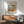 May include: Framed Studio Ghibli Ponyo poster mounted on white wall above wooden credenza with small ceramic table lamp. Green mid-century modern armchair with wooden arms sits nearby next to small round side table. Poster shows red-haired character in cozy kitchen overlooking blue ocean through large windows. Natural sunlight creates diagonal shadows across the minimalist room setting.