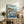 May include: Framed anime-style poster showing red-haired character surfing on large blue wave near coastal village with lighthouse, small boat in foreground, displayed on rustic wooden sideboard next to fern plant, ceramic vase, books, and beige lamp in cozy living room setting with neutral walls and brown leather furniture