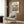 May include: Framed Studio Ghibli poster featuring Ponyo character in red dress cooking in cozy kitchen with ocean view through window, displayed on wood-paneled wall above cream upholstered armchair with geometric patterned pillow, next to brass floor lamp and dark round side table with open book