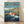 May include: Framed poster print in natural wood frame leaning against white wall on light wooden floor. Features Studio Ghibli anime art style showing red-haired character Ponyo swimming in dark blue ocean water with splash effects. Background shows coastal village houses on rocky cliffs under cloudy sky. Japanese and English text at top. Vibrant colors with teal, blue, coral and green tones in detailed illustration style.
