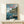 May include: Framed Studio Ghibli Ponyo poster displayed on light wood shelf against white wall. Artwork shows anime-style coastal scene with boy sitting on wooden dock, red-haired girl swimming in blue water, and traditional Japanese buildings on clifftop. Small white gift box with twine sits in front of frame. Natural lighting creates soft shadows on minimalist home decor setting.