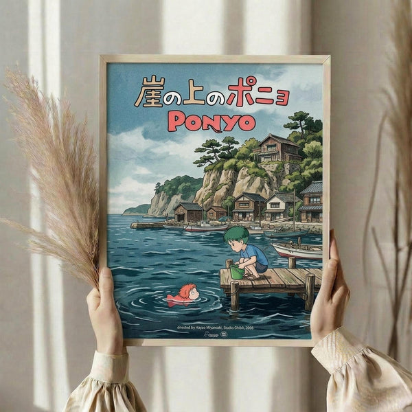 May include: Hands holding a framed Studio Ghibli Ponyo movie poster in a bright room with dried pampas grass. The artwork shows a coastal Japanese village with traditional houses on cliffs, blue water, a green-haired boy on a wooden dock, and a small red character in the water. Features Japanese text and "PONYO" in red letters against a cloudy sky background.
