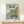 May include: Framed Studio Ghibli Ponyo poster displayed on light wood shelf against white wall. The colorful illustrated print shows a red-haired girl in orange dress standing on rocky coastline with crashing waves, clifftop village in background. Japanese and English text at top reads "A magical journey begins where the sea meets the shore." Natural wood frame, small white gift box with twine sits in front. Soft natural lighting creates gentle shadows on minimalist display.