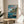 May include: Framed Ponyo movie poster in thin wooden frame displayed on round wooden side table next to beige upholstered chair. Poster features blue ocean scene with red-haired fish character swimming near wooden dock where boy sits. Japanese text at top in red and black lettering. Coastal village with traditional houses and cliffs visible in background. Soft natural lighting creates shadows across white wall. Green plant leaves partially visible in corner.
