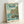 May include: Framed Ponyo movie poster in light wood frame leaning against white wall. Features anime girl with orange hair in red dress standing on rocky outcrop with ocean waves splashing around. Coastal village with houses on green cliffs in background under soft blue-green sky. Japanese text at top with "PONYO" in large dark letters and tagline below. Studio Ghibli artwork in watercolor illustration style with vintage poster aesthetic.
