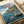 May include: Vintage-style poster print featuring Studio Ghibli's Ponyo anime artwork displayed on white paper with rolled paper tube nearby. Shows coastal Japanese village scene with green-haired boy on wooden dock reaching toward red-haired girl swimming in blue ocean water. Traditional houses built into rocky cliffs with lush trees in background under cloudy sky. Japanese text at top, director credit at bottom.