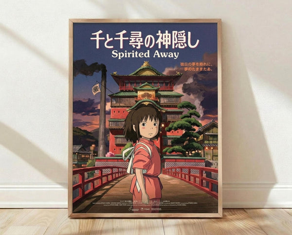 May include: Framed poster of animated girl with dark hair in pink kimono standing on red wooden bridge. Behind her is multi-story traditional Japanese bathhouse with green roofs under purple twilight sky. Poster has Japanese text at top with "Spirited Away" underneath. Wooden frame sits on light wood floor against white wall with natural lighting creating soft shadows.
