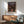May include: Framed Spirited Away movie poster hanging on white wall above wooden credenza. Poster shows young girl in pink dress on traditional Japanese street with lanterns and buildings. Mid-century modern room with mint green upholstered chair, white ceramic lamp, and natural sunlight casting shadows across the scene.