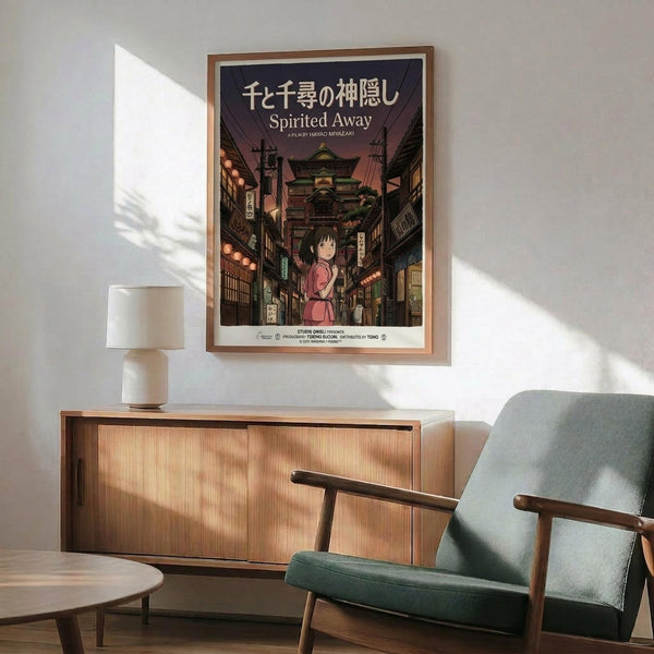 May include: Framed Spirited Away movie poster hanging on white wall above wooden credenza. Poster shows young girl in pink dress on traditional Japanese street with lanterns and buildings. Mid-century modern room with mint green upholstered chair, white ceramic lamp, and natural sunlight casting shadows across the scene.