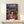 May include: Framed Spirited Away movie poster displayed on white wooden shelf next to small wrapped gift box. Features anime girl character Chihiro in pink kimono standing on red bridge with traditional Japanese bathhouse architecture in background. Poster has Japanese text at top with English "Spirited Away" subtitle. Warm wooden frame against cream wall with natural lighting creating soft shadows.