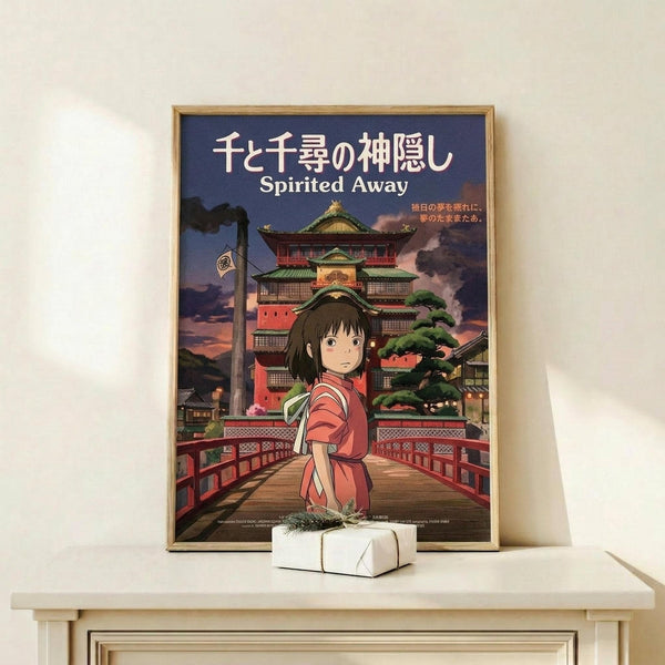 May include: Framed Spirited Away movie poster displayed on white wooden shelf next to small wrapped gift box. Features anime girl character Chihiro in pink kimono standing on red bridge with traditional Japanese bathhouse architecture in background. Poster has Japanese text at top with English "Spirited Away" subtitle. Warm wooden frame against cream wall with natural lighting creating soft shadows.
