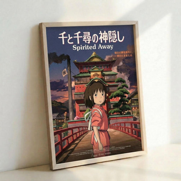 May include: Framed Spirited Away movie poster leaning against white wall corner. Features anime girl with brown hair in pink and red traditional outfit standing on wooden bridge. Multi-story green-roofed bathhouse building in background with twilight purple sky. Japanese text at top with "Spirited Away" English subtitle. Light wooden frame, vertical orientation.