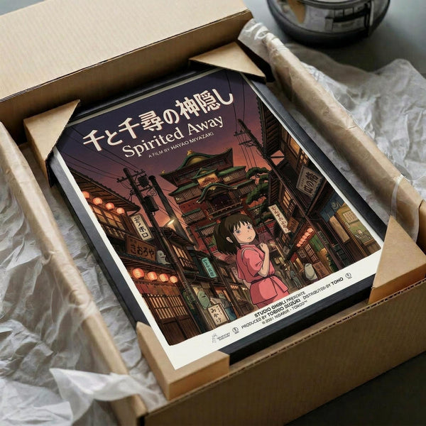 May include: Framed Spirited Away movie poster in cardboard shipping box with protective wrapping. The poster shows Chihiro, a young girl in pink outfit, standing among traditional Japanese buildings with colorful lanterns and signs. The artwork features rich purples, warm lighting, and detailed architectural elements in Studio Ghibli's distinctive animation style. Japanese and English text visible at top.