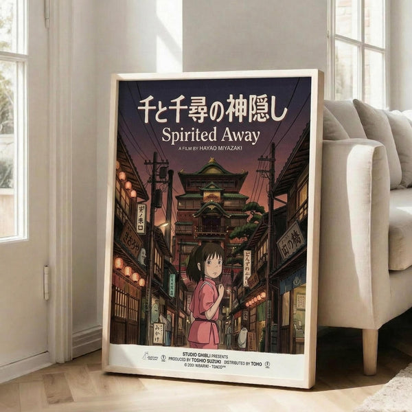 May include: Framed Spirited Away movie poster leaning against white wall next to beige sofa in bright room with tall windows. Poster shows young girl in pink dress standing on traditional Japanese street with lanterns, wooden buildings, and purple twilight sky. Japanese and English text at top. Light wooden frame, approximately 2-3 feet tall.