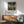 May include: Framed Spirited Away movie poster hanging on white wall above wooden credenza with white ceramic lamp. Green upholstered mid-century modern armchair with wooden frame sits beside credenza. Poster shows anime character Chihiro in marketplace scene with Japanese and English text. Natural sunlight creates shadows across the minimalist room setting.