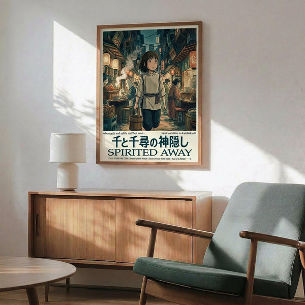 May include: Framed Spirited Away movie poster hanging on white wall above wooden credenza with white ceramic lamp. Green upholstered mid-century modern armchair with wooden frame sits beside credenza. Poster shows anime character Chihiro in marketplace scene with Japanese and English text. Natural sunlight creates shadows across the minimalist room setting.