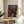 May include: Framed Spirited Away movie poster with Japanese text and English title, featuring young girl Chihiro in pink shirt standing on traditional Japanese street with lanterns and multi-story bathhouse building. Wooden frame sits on round light wood side table next to beige upholstered chair and plant leaves. Soft natural lighting creates shadows on cream wall.