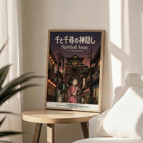 May include: Framed Spirited Away movie poster with Japanese text and English title, featuring young girl Chihiro in pink shirt standing on traditional Japanese street with lanterns and multi-story bathhouse building. Wooden frame sits on round light wood side table next to beige upholstered chair and plant leaves. Soft natural lighting creates shadows on cream wall.