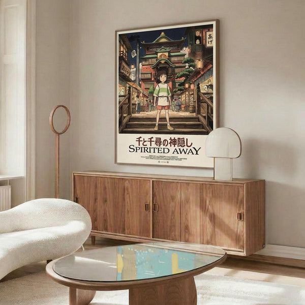 May include: Framed Spirited Away movie poster hanging above wooden credenza in modern living room with white walls, cream chair, glass coffee table, and warm lighting. Poster shows young girl character standing before colorful Japanese-style buildings and traditional architecture in anime art style.