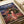 May include: Studio Ghibli Spirited Away poster print featuring Chihiro in red and white striped outfit standing on wooden deck before ornate multi-story bathhouse with traditional Japanese architecture. Rich colors include deep blues, warm reds, and golden lighting. Japanese and English text at top. Print shown on white surface with rolled papers nearby, displaying vibrant anime art style with detailed architectural elements and evening sky backdrop.