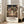 May include: Framed Spirited Away movie poster leaning against white wall next to beige sofa in bright living room. Poster shows young girl in green and white outfit standing on wooden bridge facing ornate Japanese bathhouse with colorful lanterns and traditional architecture. Artwork features rich detail in anime style with warm lighting. Japanese and English text below illustration. Light wood frame, approximately 2-3 feet tall based on furniture scale.
