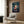 May include: Framed Spirited Away movie poster mounted on wood-paneled wall above beige upholstered armchair with geometric pillow. Poster features anime girl character with dark hair in red outfit against twilight sky with train and spirits. Modern living room setting with gold floor lamp and side table with open book.