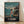 May include: Framed Studio Ghibli Spirited Away movie poster showing young girl Chihiro in red outfit standing on wooden dock beside traditional boat filled with shadowy passengers. Turquoise ocean waters stretch toward distant lighthouse and pagoda under cloudy twilight sky. Japanese text at top, English title at bottom. Natural wood frame sits on light wooden floor against white wall with soft shadows.