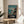 May include: Framed Studio Ghibli Spirited Away movie poster displayed on round wooden side table next to beige upholstered chair. Poster shows young girl in red outfit stepping from wooden dock onto traditional boat, with ocean and mystical buildings in background. Natural lighting creates soft shadows across white wall and plant leaves visible in corner.