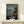 May include: Framed Studio Ghibli Spirited Away movie poster displayed on light wood shelf against cream wall. Poster shows young girl in red outfit running on wooden dock toward ferry boat, with misty ocean and mountains in background. Japanese text and "Spirited Away" title at bottom. Small white wrapped gift box sits on shelf beside the wooden-framed artwork.