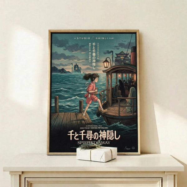 May include: Framed Studio Ghibli Spirited Away movie poster displayed on light wood shelf against cream wall. Poster shows young girl in red outfit running on wooden dock toward ferry boat, with misty ocean and mountains in background. Japanese text and "Spirited Away" title at bottom. Small white wrapped gift box sits on shelf beside the wooden-framed artwork.
