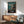 May include: Framed Studio Ghibli Spirited Away movie poster mounted on white wall above wooden sideboard. Poster shows young girl on wooden dock near traditional boat with pagoda in background, featuring teal water and cloudy sky. Mid-century modern room setting with gray upholstered chair, round coffee table, and ceramic table lamp casting natural shadows.