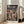 May include: A framed Studio Ghibli Spirited Away movie poster displayed in a bright living room corner beside a beige sectional sofa. The vintage-style poster features the red-haired protagonist Chihiro in traditional clothing against a fantastical Japanese bathhouse backdrop with ornate multi-story buildings, bridges, and mystical creatures. Japanese and English text overlay the dramatic cloudy scene in rich greens, browns, and golden tones within a natural wood frame.
