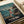 May include: Studio Ghibli Spirited Away movie poster print displayed on white paper against cream colored surface with rolled papers nearby. Poster features animated girl in red outfit standing on wooden dock beside traditional boat on water at dusk. Japanese and English text at bottom. Vintage-style illustration with teal sky and warm lighting from boat windows.