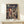 May include: Framed Studio Ghibli Spirited Away movie poster displayed on light wood shelf against cream wall. Poster shows young girl with reddish-brown hair in burgundy outfit standing before traditional Japanese bathhouse with ornate green rooftops and bustling crowd below. Japanese and English text visible. Small white wrapped gift sits on shelf beside the thin natural wood frame.