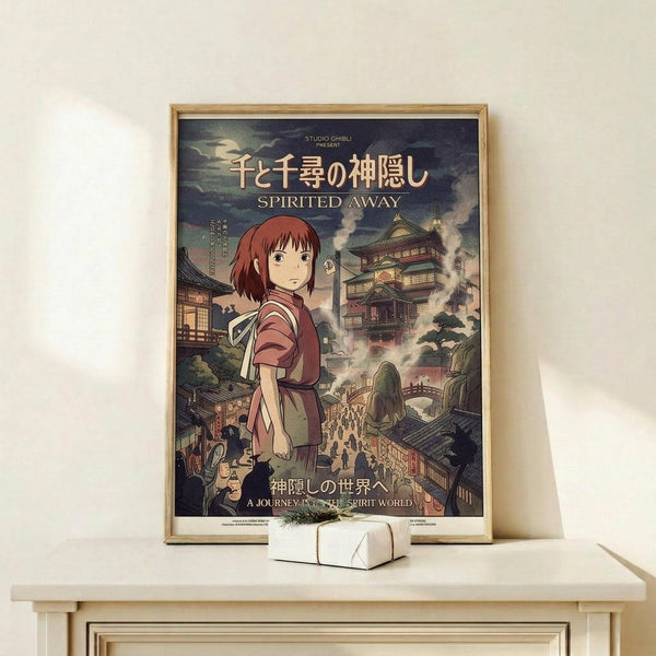 May include: Framed Studio Ghibli Spirited Away movie poster displayed on light wood shelf against cream wall. Poster shows young girl with reddish-brown hair in burgundy outfit standing before traditional Japanese bathhouse with ornate green rooftops and bustling crowd below. Japanese and English text visible. Small white wrapped gift sits on shelf beside the thin natural wood frame.