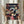 May include: Hands holding a framed Spirited Away movie poster featuring the main character Chihiro in pink shirt at center, surrounded by colorful animated characters including No-Face, dragon, and bathhouse workers against a warm lit traditional Japanese building backdrop. Golden wooden frame, neutral beige clothing and dried pampas grass visible in soft natural lighting.