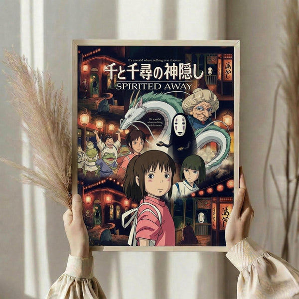 May include: Hands holding a framed Spirited Away movie poster featuring the main character Chihiro in pink shirt at center, surrounded by colorful animated characters including No-Face, dragon, and bathhouse workers against a warm lit traditional Japanese building backdrop. Golden wooden frame, neutral beige clothing and dried pampas grass visible in soft natural lighting.