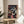 May include: Framed Studio Ghibli Spirited Away anime poster displayed on a light wood round side table next to a beige upholstered chair. The colorful poster features the main character Chihiro in a red shirt surrounded by various magical characters including No-Face, Haku the dragon, and other spirits from the bathhouse. Japanese text appears at the top with "SPIRITED AWAY" in English below. Natural sunlight streams across the minimalist room setting with a plant visible in the corner.