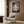 May include: Framed Spirited Away movie poster hanging on wood-paneled wall above beige upholstered armchair with geometric patterned pillow. Poster shows anime characters including young girl and mysterious black figure beneath traditional Japanese torii gate, surrounded by fantastical creatures in muted colors. Modern living room setting with brass floor lamp, dark round side table with open book, and white curtains.