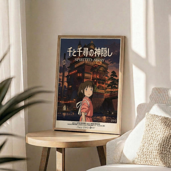 May include: Framed Spirited Away movie poster displayed on wooden round side table next to beige chair with knitted throw pillow. Poster shows anime girl in red outfit against twilight bathhouse scene with Japanese text. Natural lighting creates soft shadows on white wall with plant leaves visible.