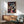 May include: Framed Spirited Away movie poster hanging on white wall above wooden credenza with lamp. Features colorful anime artwork showing young girl Chihiro in pink shirt surrounded by magical characters including No-Face, dragon, and bathhouse scenes. Modern living room setting with teal mid-century chair and round coffee table. Natural lighting creates geometric shadows on wall.