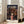 May include: Framed Spirited Away movie poster leaning against beige sofa in bright living room with white walls and large windows. Poster features anime girl in pink outfit standing before ornate Japanese bathhouse at twilight with warm golden lighting reflecting on water. Natural wood frame, cream matting, positioned on light wooden floors next to modern furniture.