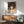 May include: Framed Spirited Away movie poster featuring Chihiro, a young girl with dark hair surrounded by white spirits, displayed above a wooden sideboard with ceramic lamp in a modern living room with teal upholstered chair and natural lighting creating geometric shadows on white walls.