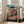 May include: Framed anime poster leaning against white wall next to beige sofa in bright living room. Artwork shows young girl in pink outfit jumping over red bridge with traditional Japanese bathhouse building in background. Colorful illustration features turquoise sky, green trees, and Japanese text at bottom. White wooden frame on light hardwood floors with natural window lighting.