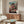 May include: Framed Studio Ghibli anime poster displayed above wooden sideboard in modern living room. Artwork shows young girl in pink outfit leaping across traditional Japanese buildings and red bridge, with Japanese text at bottom. Room features cream upholstered chair, glass coffee table, and minimalist decor in neutral tones.